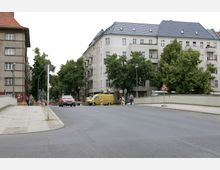 Straßenszene in einem städtischen Wohngebiet mit mehrstöckigen Wohnhäusern. Auf der Straße stehen geparkte Autos, ein gelber Lieferwagen und Bauabsperrungen; Fußgänger benutzen den Bürgersteig, und Bäume säumen den Bereich.