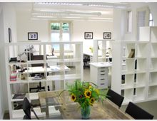 Helles Büro mit mehreren Arbeitsplätzen, weißen Regalen als Raumtrenner und großen Fenstern, die natürliches Licht hereinlassen. Im Vordergrund steht ein Tisch aus Holz mit einem Blumenstrauß aus Sonnenblumen.