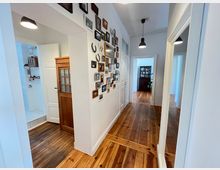 Ein Flur in einer Wohnung mit Holzdielenboden, weißen Wänden und schwarzer Hängebeleuchtung. An der rechten Wand hängen zahlreiche gerahmte Fotos, links ist eine Tür geöffnet und zeigt ein weiß gefliestes Badezimmer, während im Hintergrund ein Raum mit einer Holzanrichte sichtbar ist.