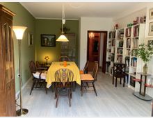 Esszimmer in einem wohnlichen Stil mit einem Esstisch, der mit einer gelben Tischdecke bedeckt ist, umgeben von Holzstühlen. Links steht ein hoher Schrank vor einer grün gestrichenen Wand, rechts befinden sich weiße Bücherregale, und im Hintergrund führt eine Tür zu einem anderen Raum mit einem Kronleuchter.