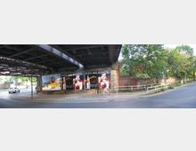 Brücke mit Ziegelmauerwerk, unter der Plakatwerbung angebracht ist. Rechts verläuft ein Gehweg mit Geländer entlang einer alten Ziegelmauer, dahinter befinden sich Bäume und Vegetation. Eine Straße führt unter der Brücke hindurch.