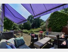 Gemütliche Terrasse mit Sitzmöbeln und Dekokissen, überdeckt von lilafarbenen Markisen. Im Hintergrund ein Garten mit grünen Pflanzen und ein Backsteingebäude mit von Efeu bewachsener Fassade.