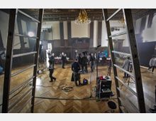 Ein großer, dekorierter Saal mit Holzparkettboden, hohen Säulen und einer verzierten Decke. Im Vordergrund befinden sich Filmgeräte wie Mikrofone, Scheinwerfer und Kabel, während im Hintergrund Menschen an Tischen sitzen und stehen.