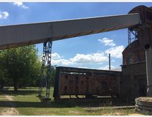 Industriegelände mit einem alten Backsteingebäude, einem angrenzenden verrosteten Schornstein und einem erhöhten, metallverkleideten Förderband, das auf Stahlgerüsten ruht. Im Hintergrund sind grüne Bäume und ein blauer Himmel mit weißen Wolken zu sehen.