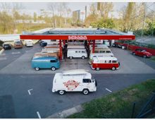 Eine alte Tankstelle mit mehreren geparkten VW-Bussen in verschiedenen Farben, darunter rote, weiße und blaue Modelle. Im Vordergrund ein weißer VW-Bus mit der Aufschrift „Bulli Tanke Berlin“, im Hintergrund Bäume und moderne Gebäude.