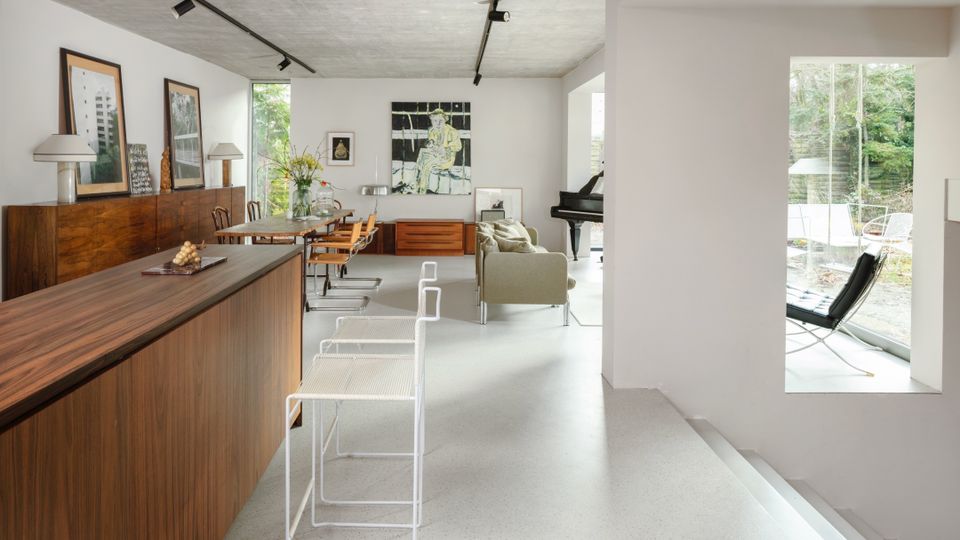 Open-plan modern living and dining area with a long wood cabinet and dining table, white walls, and a raw concrete ceiling with track lighting. Large floor-to-ceiling windows bring in garden views, with a sitting area, a grand piano, and modern chairs arranged throughout.