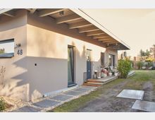 Moderne eingeschossige Wohnhausfassade mit flachem Pultdach. Ein gepflasterter Weg führt zur Haustür, die von großen Glasfenstern umgeben ist, und eine Terrasse mit Töpfen und Möbeln befindet sich an der Seite; im Hintergrund sind Gartenbereiche und vereinzelte Sträucher sichtbar.