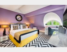 Schlafzimmer mit lila Wandfarbe und gemustertem Teppichboden im Zickzack-Design. Das Zimmer verfügt über ein Doppelbett mit verziertem Kopfteil, zwei Nachttische mit Lampen und einen Schreibtisch vor einem großen, runden Fenster mit Blick ins Grüne.