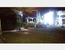 Ein beleuchteter Garten bei Nacht mit einem kleinen weißen Pavillon, umgeben von Sträuchern und Bäumen. Rechts im Bild ist eine weiß angestrahlte Hausfassade mit geöffneten Fenstern und Beleuchtungsequipment auf Stativen zu sehen.