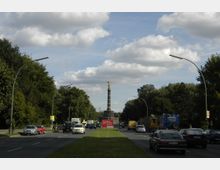Straße mit mehreren Fahrbahnen im Tiergarten in Berlin, gesäumt von Bäumen und modernen Straßenlaternen, im Hintergrund die Siegessäule mit der goldenen Figur auf der Spitze. Autos fahren in beide Richtungen, und eine grasbewachsene Mittelspur trennt die Fahrbahnen.