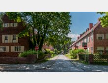 Straßenansicht in einem Wohnviertel mit gepflasterter Fahrbahn, gesäumt von roten Backsteinhäusern mit weißen Fensterläden. Große Bäume und Sträucher spenden Schatten, und an beiden Straßenseiten sind Verbotsschilder sichtbar.