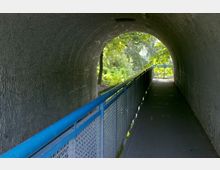 Ein schmaler Tunnel mit Betonwänden, der von Tageslicht am Ende erhellt wird. Ein Gehweg mit einem blauen Geländer verläuft durch den Tunnel und führt in eine grüne, baumbewachsene Umgebung.