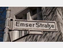 Ein Straßenschild mit der Aufschrift "Emser Straße", befestigt an einer grauen Metallstange. Im Hintergrund ist die Fassade eines Gebäudes mit weißen Fensterrahmen zu sehen.