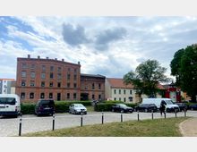 Ein mehrstöckiges Backsteingebäude mit großen Fensterreihen steht neben einem kleinen, hellen Gebäude mit roten Ziegeldächern. Im Vordergrund befindet sich ein Parkplatz mit mehreren geparkten Autos und ein Gehweg, dahinter einige Bäume und ein bewölkter Himmel.