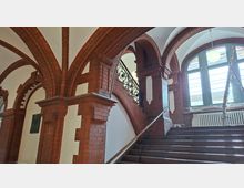 Innenraum eines historischen Treppenhauses mit roten Backsteinbögen und weißem Gewölbe, einer breiten Steintreppe mit Geländer sowie dekorativem Schmiedeeisengitter. Großes Rundbogenfenster mit Heizkörper darunter; eine Leiter steht am Fensterbereich.