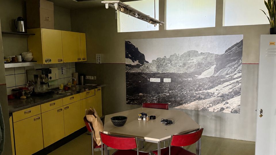 Blick in eine Gemeinschaftsküche mit gelben Schrankfronten, Edelstahl-Arbeitsbereich und einem sechseckigen Tisch mit roten Stühlen. An der Wand hängt ein großformatiges Schwarzweiß-Foto einer Berglandschaft, darüber befinden sich hohe Fenster.