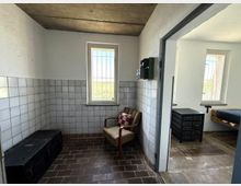 Ein einfacher Innenraum mit gefliesten Wänden, braunem Ziegelboden und zwei Fenstern mit Gitterstäben. Links steht eine schwarze Truhe, in der Mitte ein gepolsterter Stuhl mit kariertem Kissen, und im angrenzenden Raum sind eine Metall-Kommode und ein Holzbett erkennbar.