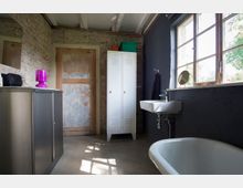 Badezimmer in einem rustikalen Stil mit unverputzter Ziegelwand, einer hölzernen Tür und sichtbarer Balkendecke. Der Raum enthält eine weiße freistehende Badewanne, ein Waschbecken unter einem großen Fenster und einen weißen Metallschrank.