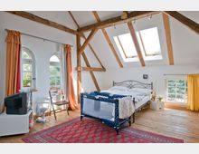 Dachzimmer mit sichtbaren Holzbalken, einem Doppelbett, einem Babyreisebett und einer Einrichtung im Landhausstil. Die weißen Wände sind mit Dachfenstern und bogenförmigen Fenstern versehen, die mit orange-karierten Vorhängen dekoriert sind.