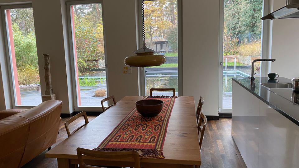 Offener Ess- und Küchenbereich mit großem Holztisch, Stühlen und einer Hängelampe; rechts eine moderne Küchenzeile mit Spüle. Mehrere bodentiefe Fenster und eine Glastür geben den Blick auf eine Terrasse und einen Garten mit Pflanzen frei.