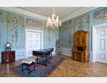 Prächtiger Raum im Rokoko-Stil mit aufwendig bemalten Wänden in Pastelltönen, verziert mit Blumen- und Goldornamenten, einer Kristallkronleuchter-Deckenlampe, dekorativen Möbeln aus Holz und einem verzierten Klavichord auf einem orientalischen Teppich.