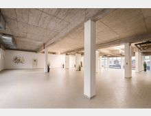 Großer, leerer Innenraum im Industrie‑Loft-Stil mit Sichtbetondecke, weißen Stützsäulen und hellem Fliesenboden. Weiße Trennwände mit einem Kunstbild, mehrere Topfpflanzen sowie große Fensterfronten zur Straße prägen den Raum.