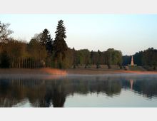 Ein ruhiger See mit Schilfgürtel im Vordergrund spiegelt die Baumreihe eines bewaldeten Ufers wider. Im Hintergrund steht ein steinerner Obelisk auf einer erhöhten, offenen Wiesenfläche, umgeben von weiteren Bäumen.