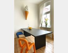 Ein heller Raum mit Holzboden, einem schwarzen Holztisch und einem markanten orangefarbenen Stuhl mit geschwungener Form. An der weißen Wand hängt eine große Eiscreme-Dekoration, und ein Fenstersims mit Gardinen zeigt Blumenvasen und Blick nach draußen.