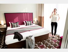 Ein modernes Hotelzimmer mit einem Doppelbett, das mit einem burgunderroten Kopfteil, grauen und rosa gestreiften Kissen sowie einer passenden Tagesdecke gestaltet ist. Im Raum steht eine Frau telefonierend neben Vorhängen, die durch ein großes Fenster Licht hereinlassen.
