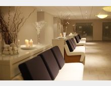 Moderner Wartebereich mit weißen Sitzbänken und dunklen Kissen entlang einer hellen Wand. Dekorative Elemente wie Kerzen, Glasvasen mit Zweigen und weiße Flusssteine stehen auf hohen, schmalen Tischen, während die Umgebung durch warmes Licht und dezente Deckenlampen beleuchtet wird.