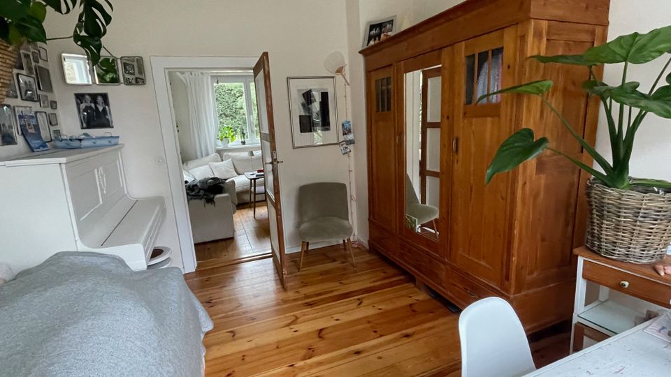 Heller Wohnraum mit Holzdielen, weißem Klavier an der linken Wand und großem Holzkleiderschrank mit Spiegeltür rechts. Eine offene Glastür führt in ein angrenzendes Wohnzimmer mit Sofa und Fenster; an den Wänden hängen Fotos und Bilder, dazu stehen Zimmerpflanzen.