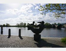 Eine Bronzeskulptur einer Meerjungfrau mit Kind steht auf einem gepflasterten Platz am Ufer eines Flusses. Im Hintergrund sind Bäume, eine Brücke und der leicht bewölkte Himmel zu sehen.
