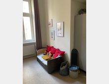 Ein Kinderzimmer mit einem kleinen Sofa, das mit roten Kissen und Stofftieren dekoriert ist. An der Wand hängen drei Bilder mit kindlichen Motiven, daneben steht ein Schrank sowie zwei Stoffkörbe und ein Skateboard.