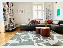 Helles, modernes Wohnzimmer mit großem L-förmigem Sofa, gemustertem Teppich und zwei runden Couchtischen aus Holz. Links stehen deckenhohe Bücherregale, hinten ein Fenster sowie Wandkunst und eine hohe Bogenlampe.