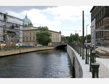 Stadtkanal mit einer steinernen Brücke im Hintergrund, flankiert von einem sanierten Gebäude mit Kuppel und einem Baugerüst links und einem historischen Gebäude rechts; im Hintergrund sind Bäume, Baukräne und parkende Autos zu sehen.
