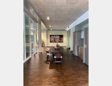 Moderner Innenraum mit einem langen Esstisch in der Mitte, umgeben von Stühlen mit unterschiedlichen Farben. Der Raum hat einen Boden aus dunklem Holz, eine graue Betondecke und Seitenwände mit Glaselementen sowie einen Durchgang zur Küche rechts. Im Hintergrund ist eine Wand mit einem bunten Blumenbild und einer dekorativen Vase sichtbar.