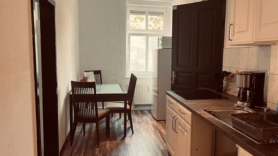 Schmale Wohnküche mit Holzoptikboden, Esstisch und vier Stühlen vor einem großen Fenster. Rechts eine weiße Küchenzeile mit Spüle und Arbeitsplatte sowie ein hoher Kühlschrank neben einer dunklen Tür.