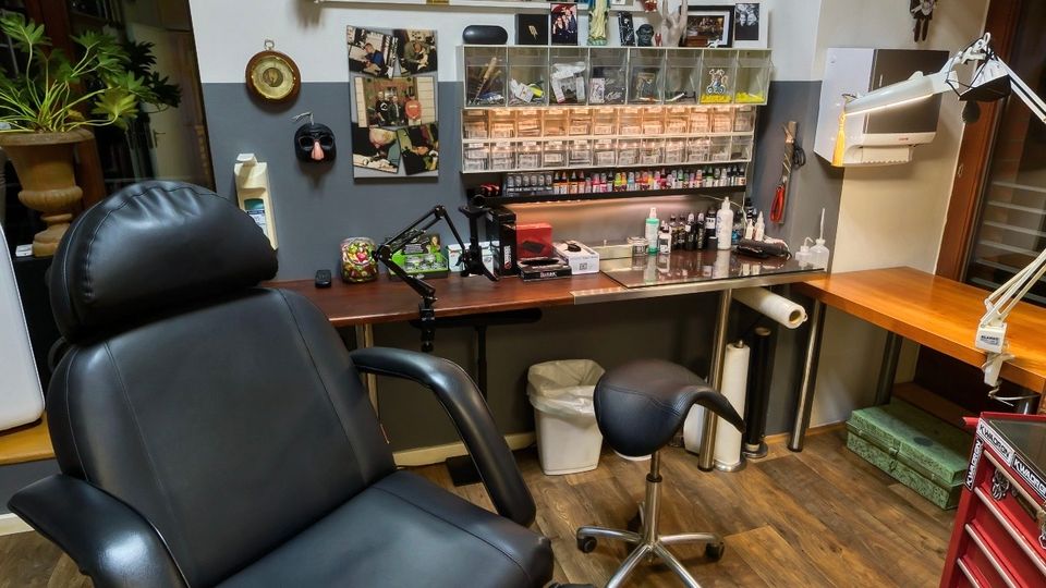Innenraum eines Tattoo-Studios mit einem großen schwarzen Liegestuhl, Holzfußboden und Arbeitsflächen mit Lampen, Geräten und farbigen Tintenfläschchen in Regalen. Die Wände sind dicht mit gerahmten Bildern, Urkunden und Dekorationen versehen, neben Fenstern und einer Werkzeugkommode.