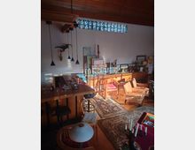 Wohnraum mit Holzbalkendecke und dekorativem Oberlicht aus buntem Glas. Der Raum enthält eine Bar mit Hockern, gemütliche Sitzmöbel, Teppiche und verschiedene Kunstgegenstände sowie Dekorationen.