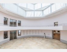 Großer, heller Innenraum eines modernen Museums mit glatten, weißen Wänden und einem achteckigen Glasdach, das den Raum mit natürlichem Licht durchflutet. An den Wänden sind Texttafeln angebracht, und durch Tür- und Fensteröffnungen sind weitere Ausstellungsräume sichtbar; der Boden besteht aus beigefarbenen Steinfliesen.