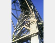 Nahaufnahme eines stählernen Brückengerüsts mit sichtbaren Streben, Treppen und Geländern, die komplexe Strukturen und Verstrebungen zeigen. Der Hintergrund ist ein klarer blauer Himmel.