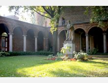 Ein Innenhof mit einer Statue im Vordergrund, umgeben von einem Arkadengang mit Backsteinwänden und Rundbögen, sowie einer Rasenfläche mit Blumenbeeten. Bäume und Sonnenlicht schaffen eine helle, ruhige Atmosphäre.