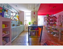 Bright, colorful open-plan kitchen and dining area with a long counter and sink beneath stained-glass style windows, a central island worktable, and wooden plank flooring. The adjoining dining space features a vivid red ceiling, mosaic-tiled table, pastel chairs, open shelving with plants, and framed wall art.