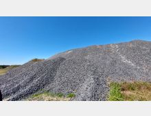 Ein großer Schotterhaufen auf einer offenen Fläche mit vereinzelten Gräsern im Vordergrund, unter einem klaren blauen Himmel.