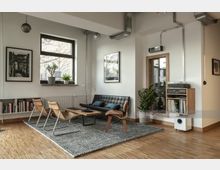 Helles Wohnzimmer mit modernem, minimalistischem Interieur, einschließlich eines schwarzen Ledersofas, Holzmöbeln und einem grauen Teppich auf Parkettboden. Große Fenster, Zimmerpflanzen, eingerahmte Wandbilder und ein Regal mit Büchern und Audiogeräten ergänzen den Raum.