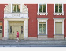 Fassade eines historischen Gebäudes aus rotem Ziegelstein mit weißen Stuckverzierungen um die Fenster und Türen. Eine gelbe Holztür mit dekorativen Eisengittern bildet den Eingang, während eine Frau in roter Kleidung den Gehweg entlangläuft.