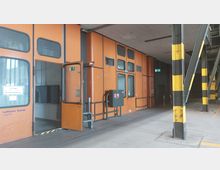 Ein Lagerbereich mit orangefarbenen Wänden, großen Glastüren und einem Schild mit der Aufschrift "Zollager – Bonded Warehouse". Im Vordergrund befinden sich metallene Geländer und gelb-schwarz markierte Stützpfeiler, die den industriellen Charakter der Umgebung unterstreichen.