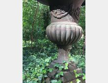 Große, verzierte Steinvase mit reliefartigen Motiven, umgeben von üppigem Efeu und dichter Vegetation im Hintergrund. Die Vase steht auf einem steinernen Sockel in einer bewachsenen Wald- oder Gartenlandschaft.