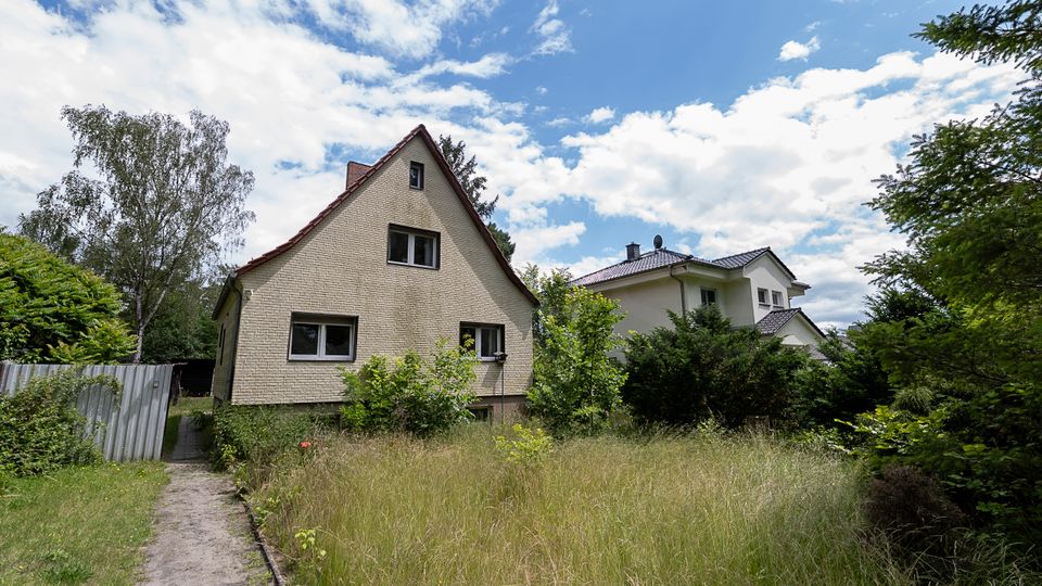 Blick auf ein freistehendes Einfamilienhaus mit steilem Satteldach und hell verklinkerter Fassade in einer grünen Wohngegend. Davor liegt ein verwilderter Garten mit hohem Gras, Büschen und einem schmalen Weg; im Hintergrund steht ein weiteres Haus unter blauem Himmel mit Wolken.
