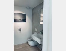 Modernes Badezimmer mit einer hängenden Toilette, einem rechteckigen Waschbecken mit Wandarmatur und einem schlichten Design. Die Wände sind weiß und grau, mit einem minimalistischen Kunstwerk an der Wand und einem Holzboden.